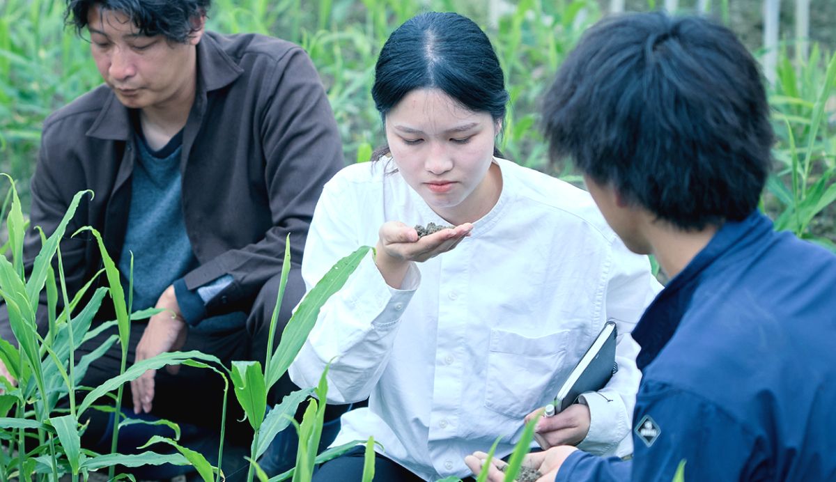 植物の前に3人がしゃがんでいる。真ん中の女性は土を手に乗せて匂いを嗅いでいる。